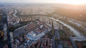 Veduta aerea di Roma al tramonto, con il fiume Tevere che attraversa la città formando curve sinuose. L'area urbana è densa di edifici residenziali e storici, con strade alberate e ampi complessi architettonici. In primo piano, un grande edificio moderno con una struttura geometrica si distingue tra i tetti rossi delle costruzioni circostanti. Sullo sfondo, la città si estende fino all'orizzonte, avvolta in una luce dorata.